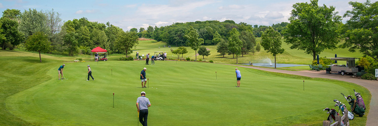 Bethany Lutheran College students are playing golf on a green course with trees, a fountain, a red canopy, and golf carts in the background.