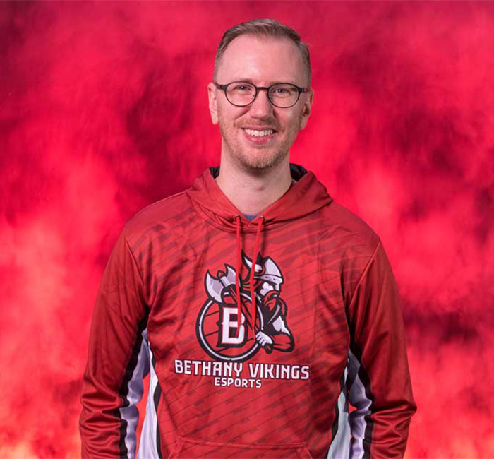 A Bethany Lutheran College student in glasses and a Vikings Esports hoodie smiles confidently before a red, smoky backdrop.