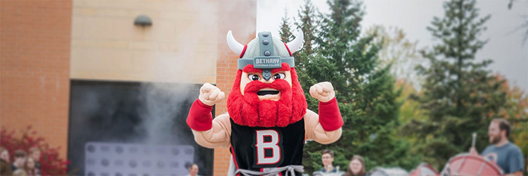 A Viking mascot with a red beard and horned helmet stands with raised fists near trees and a building at Bethany Lutheran College.