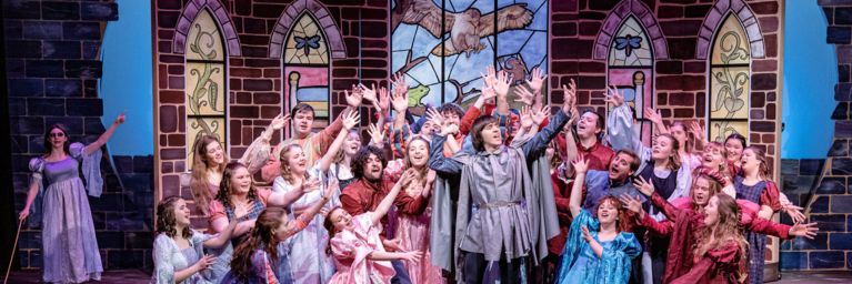 Bethany Lutheran College students in colorful period costumes sing onstage before stained glass windows and a painted backdrop, likely performing in a theatrical or musical production.