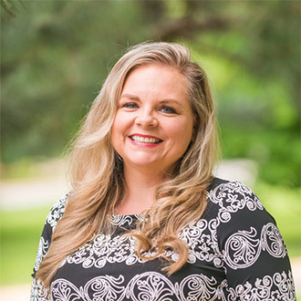 A smiling woman with long blonde hair, wearing a patterned top, stands outdoors on a Minnesota campus with greenery in the background.