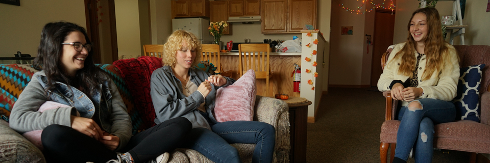 Three Bethany Lutheran College students sit and talk in a cozy Minnesota living room decorated for fall, with a kitchen visible in the background.