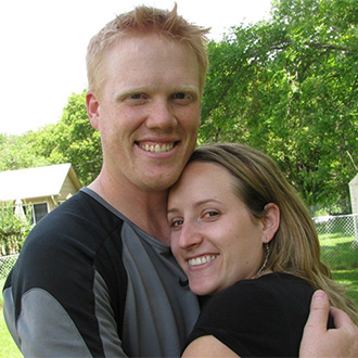 A smiling couple embraces outdoors at Bethany Lutheran College in Minnesota, with trees and houses visible in the background.