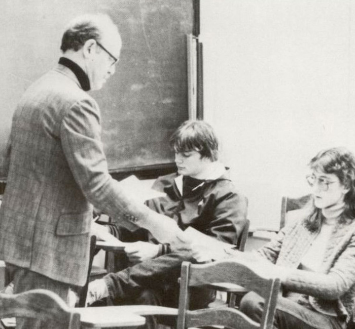 A teacher hands out papers to two Bethany Lutheran College students seated at desks in a classroom with a chalkboard in the background.