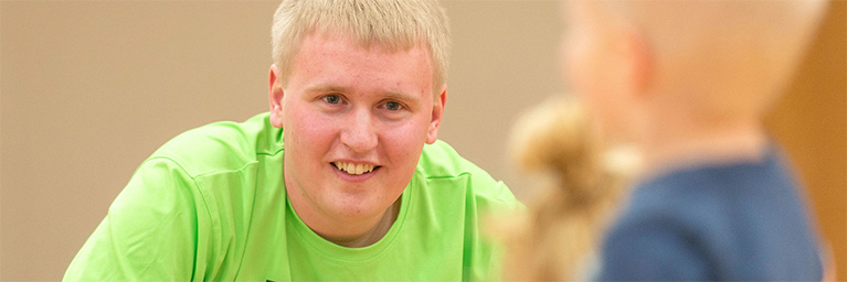 A young man in a bright green shirt smiles while interacting with a child on a Minnesota campus.