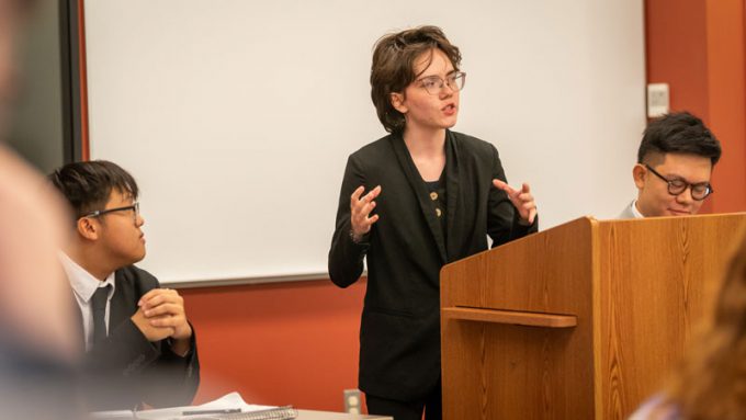 A Bethany Lutheran College student stands and speaks at a podium while two other students, also in formal attire, are seated nearby.