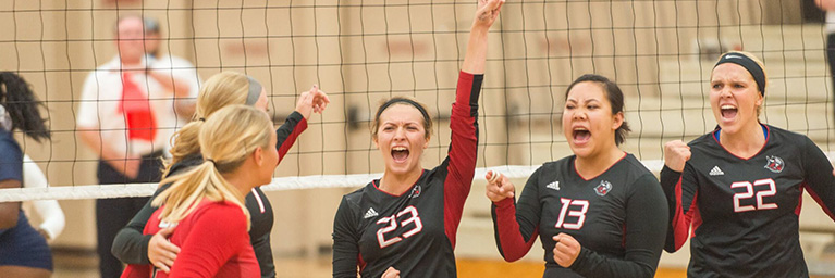 Five Bethany Lutheran College volleyball players wearing black jerseys celebrate together at the net during a match.