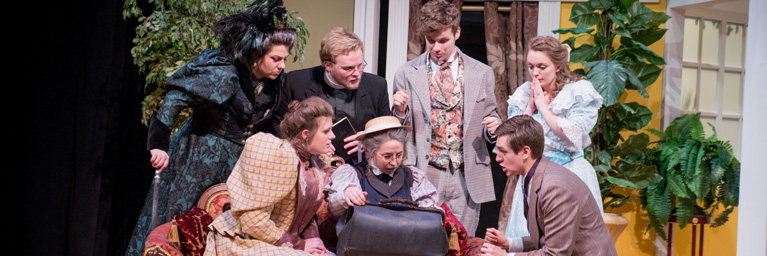 Seven actors in period costumes gather around a student with a suitcase on stage during a Bethany Lutheran College performance in Minnesota.