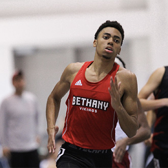 Bethany Lutheran College track athlete wearing a red Vikings jersey runs indoors, with a blurred campus community in the background.