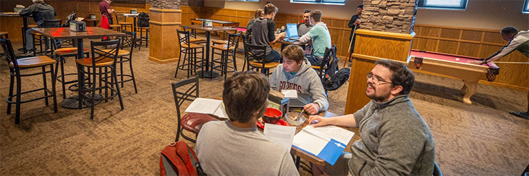 Students sit at tables in a Bethany Lutheran College lounge, studying and talking, with a pool table visible in the background.