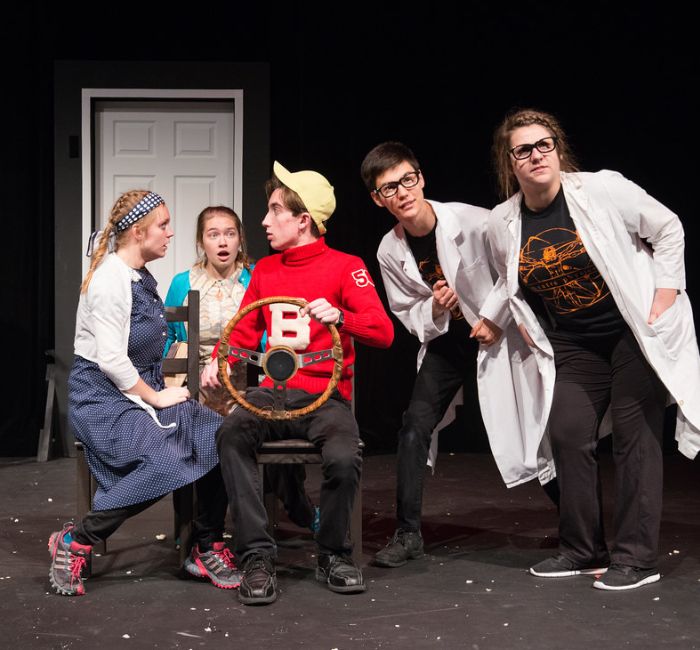 Five Bethany Lutheran College students perform on stage in Mankato; one student holds a steering wheel while the others act as scientists and a farmer, engaging in a lively scene.