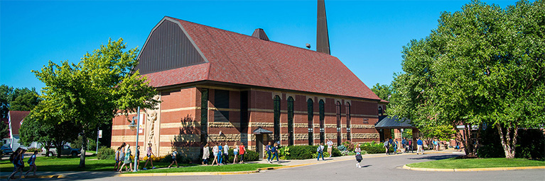 Students walk near the Trinity Chapel building with a red roof, surrounded by tall trees and set against a clear blue sky on a sunny day.