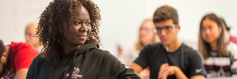 A woman in a black hoodie sits in a Mankato classroom, smiling, with students engaged in activities visible in the background.