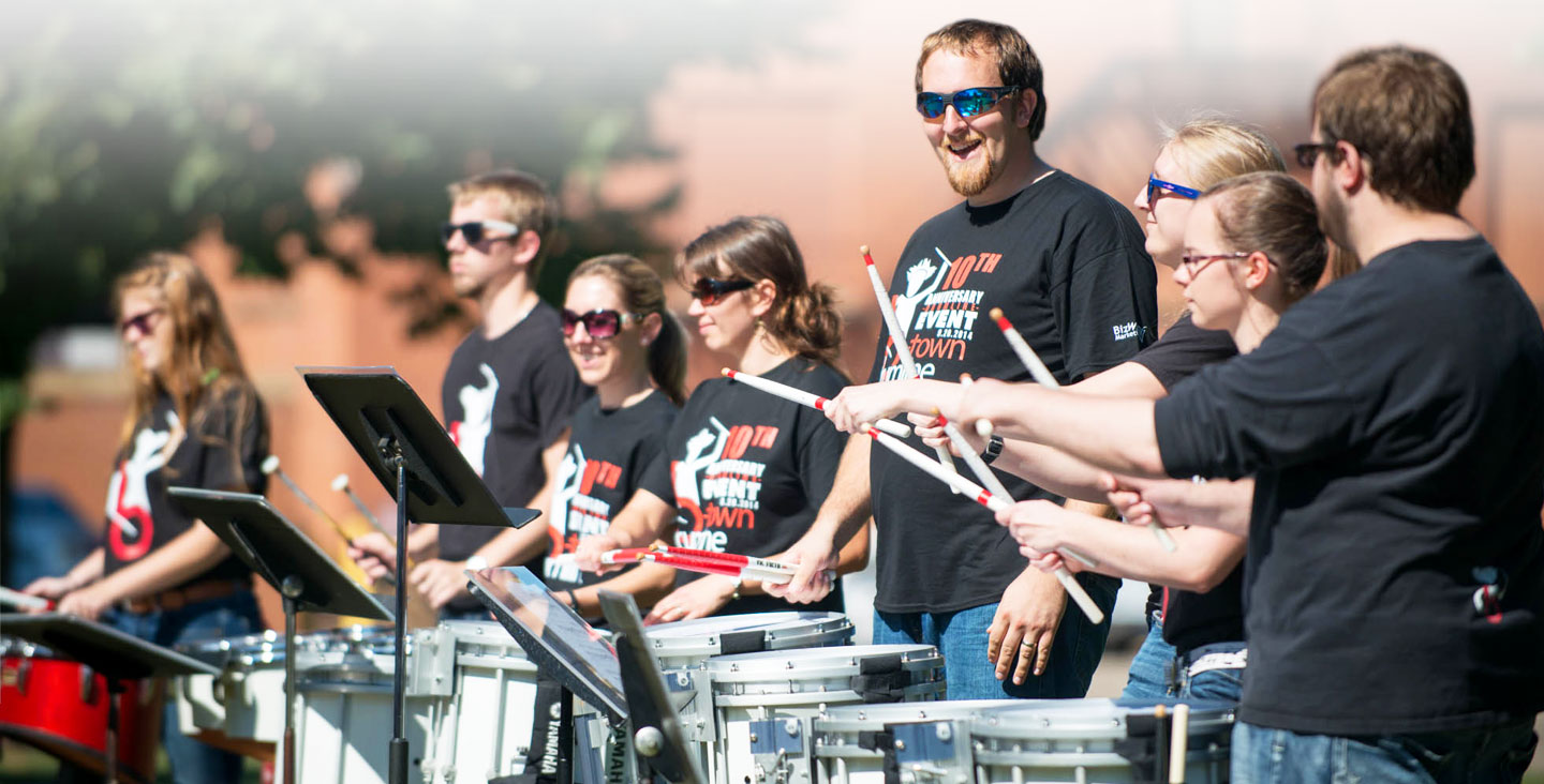 A group of people wearing sunglasses stands in a row, smiling and playing drums outdoors on a Minnesota campus.