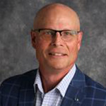 A bald man wearing glasses, a checkered shirt, and a blue plaid blazer poses in front of a gray backdrop at Bethany Lutheran College.