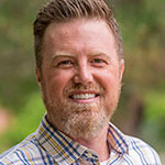 Chris Bollig, Adjunct Faculty for the Clinical Mental Health Counseling – Graduate Programs Department, stands outdoors with greenery in the background.