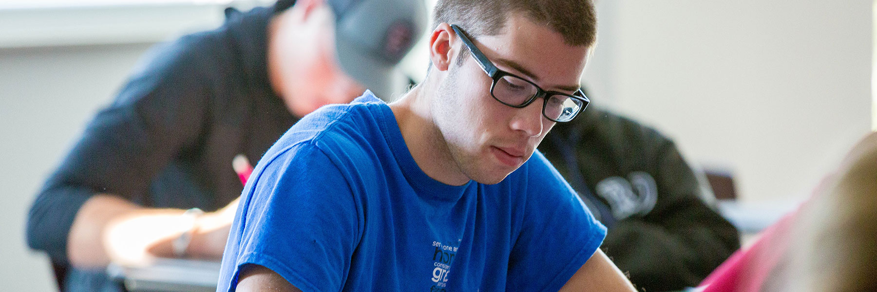 Young man wearing glasses and a blue shirt writes at a desk, while other students in the classroom appear engaged.