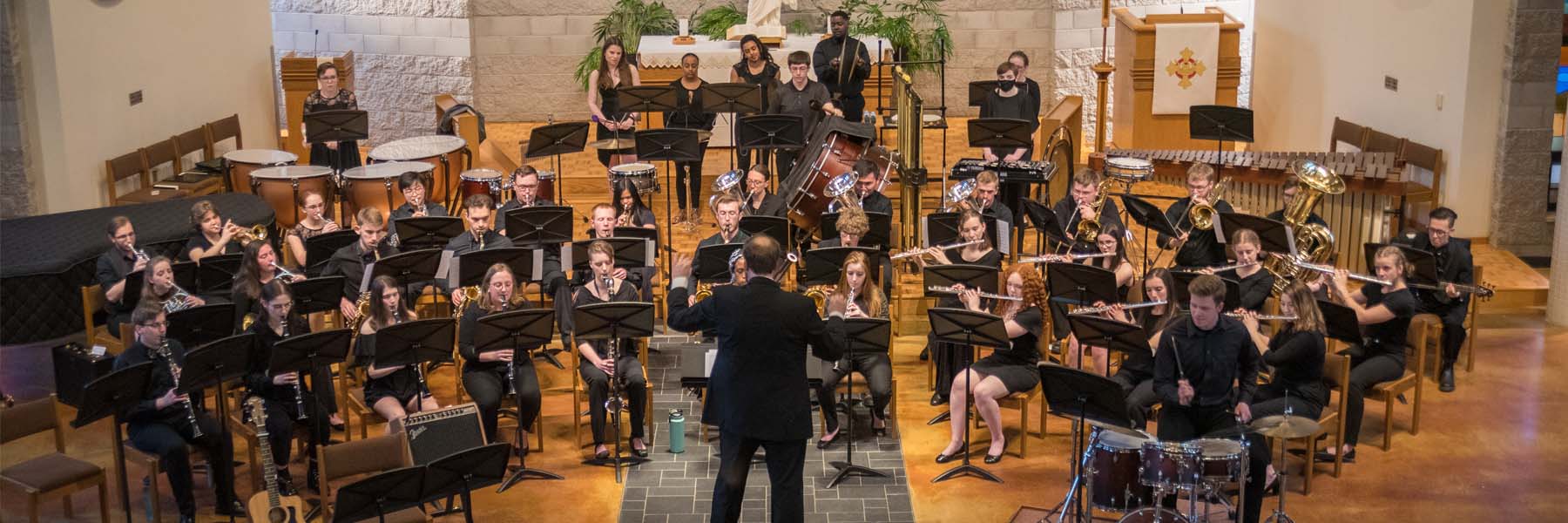 A conductor leads a large ensemble of musicians playing various instruments for the campus community in a church setting.