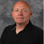 Derick Lyngholm, Associate Professor for the Communication Department and Head Coach Men's Soccer for the Athletics Department, poses in front of a gray background, representing the Bethany Lutheran College campus community.