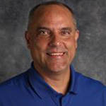 Eric Woller, Professor for the Chemistry Department, poses in front of a gray background.