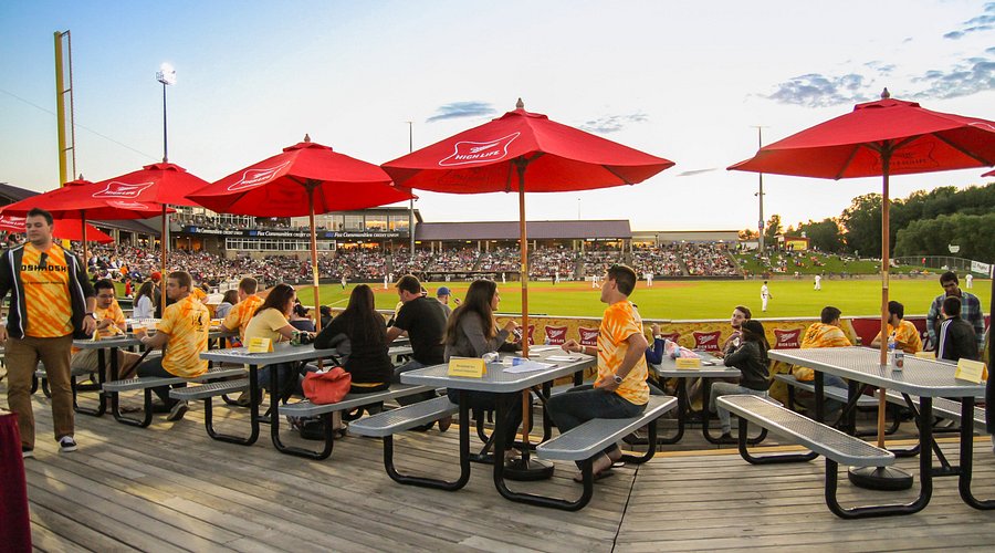 Bethany Lutheran College students sit at picnic tables with red umbrellas, watching a baseball game at a Minnesota stadium during sunset.