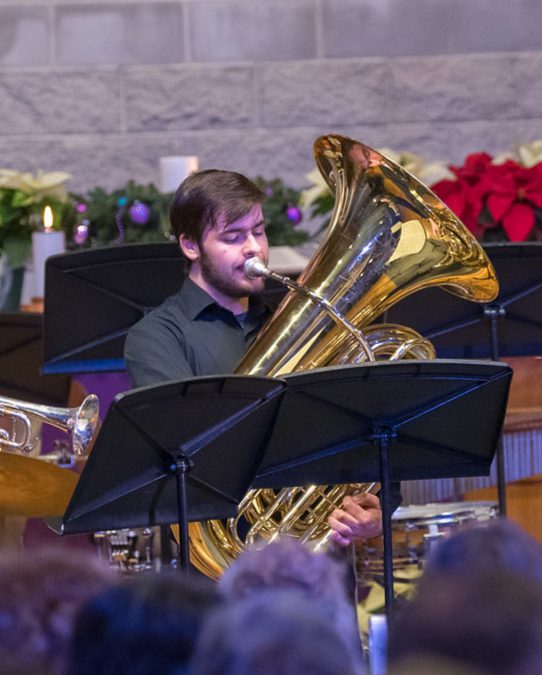A man plays a tuba during a concert at Bethany Lutheran College, with music stands and holiday decorations in the background.