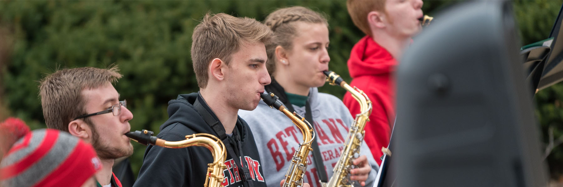 Four young people play saxophones outdoors in Mankato, wearing jackets. Trees and campus buildings are visible in the background.