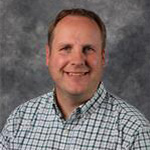 John Gunderson, Associate Professor for the Languages Department and Study Abroad Coordinator for the Academic Affairs Department, poses in front of a gray mottled background.