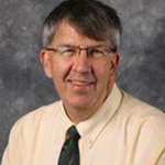 John Sehloff, Director of the Information Technology Services Department and Online Instructor for the Computer Science Department, smiles in front of a gray mottled background.