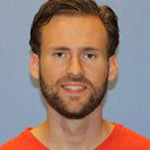 Johnathan Moeller, Lessons Instructor for the Music Department, smiles in front of a blue background.
