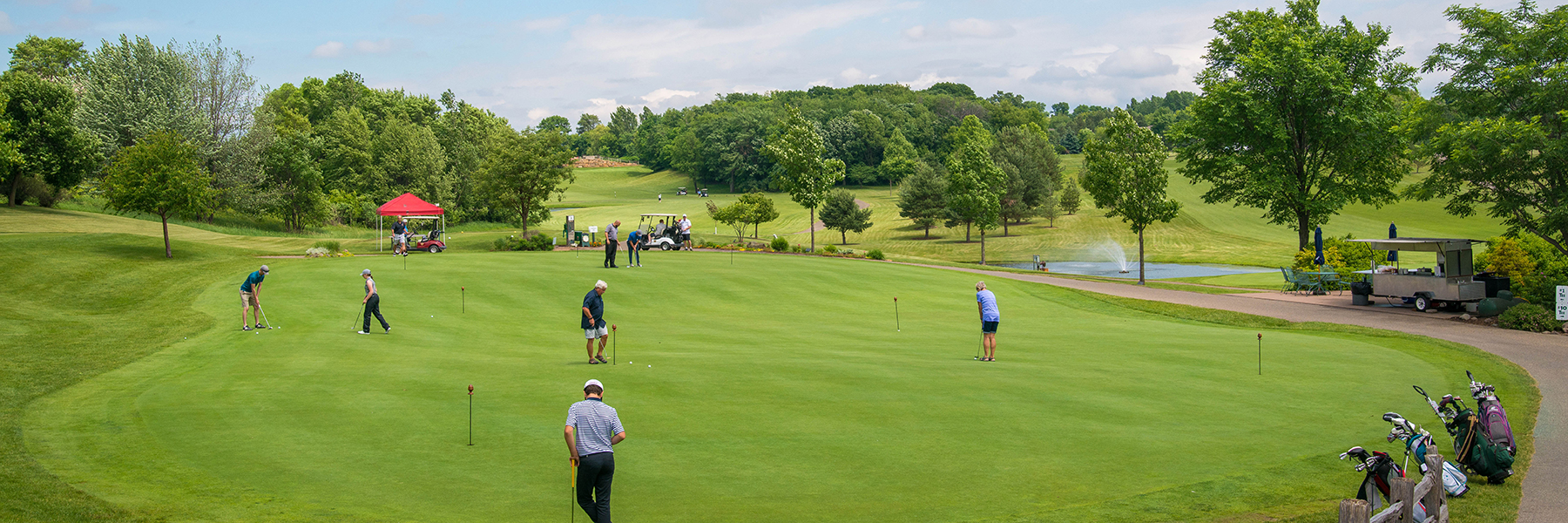Golfers putting on a large green in Mankato, with trees, golf carts, a red tent, and a fountain nearby.