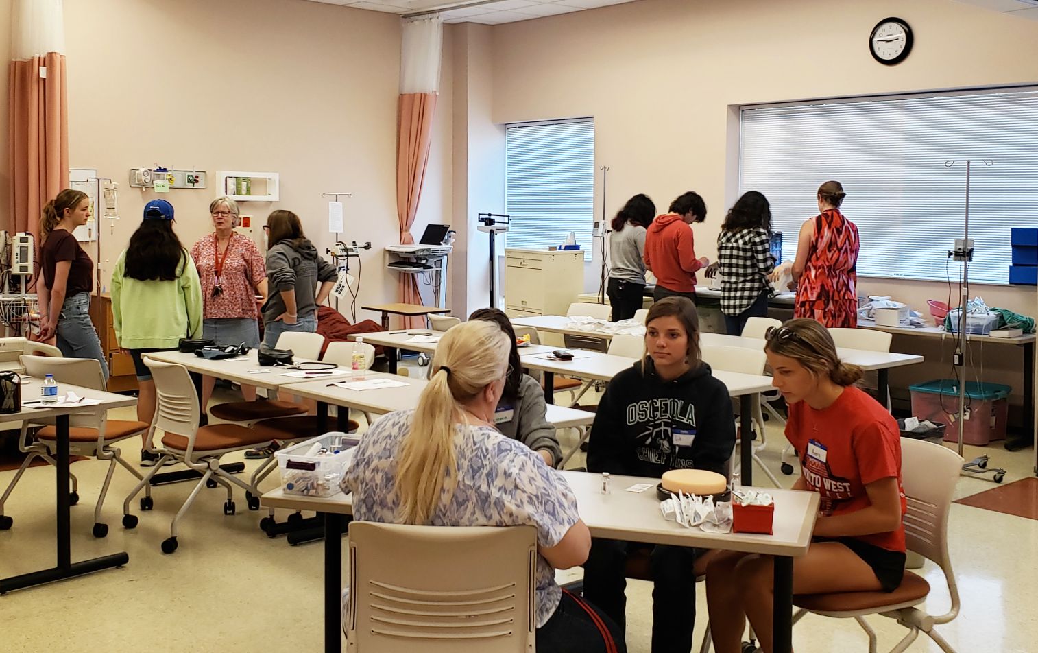 Students at Bethany Lutheran College engage in medical skills practice at workstations, with teachers and medical equipment visible.