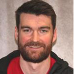 Ryan Nichols, Head Coach Women's Soccer, Weight Room Monitor, and Game Management for Athletics Department, stands smiling in front of a gray backdrop at Bethany Lutheran College.