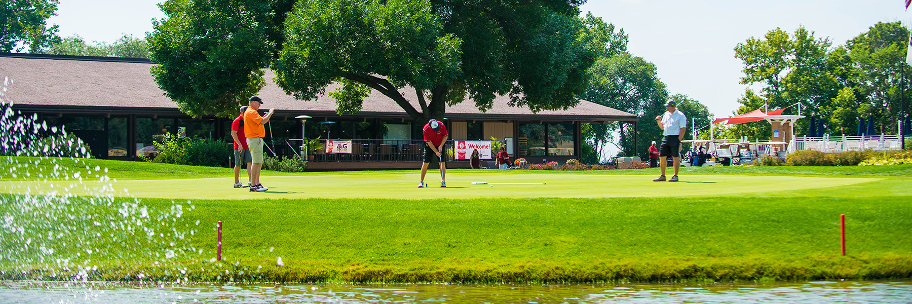 Three Bethany Lutheran College students are playing golf on a green near a pond, with the college clubhouse and Minnesota trees visible in the background.
