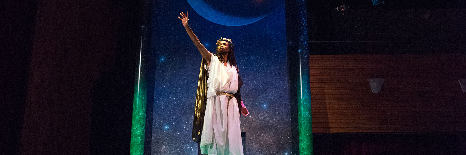 A person wearing a white robe stands on stage at Bethany Lutheran College, with a starry backdrop and crescent moon behind them.