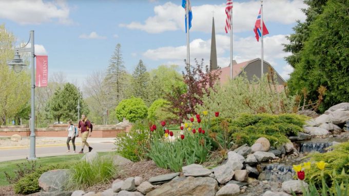 Two people walk along a campus path in Mankato, MN, near red tulips, rocks, and flags on a sunny day with scattered clouds.