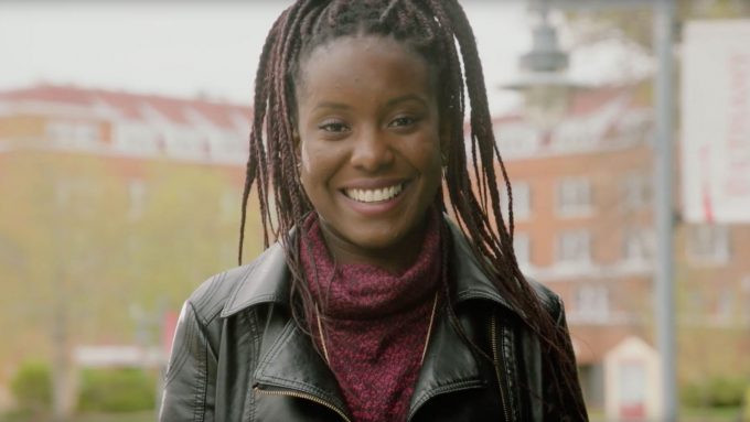 A person with long braided hair smiles outdoors on a campus, wearing a black leather jacket and a maroon sweater. Buildings are visible in the background.