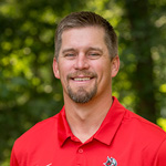 CJ Siewert, Director of Sports Information with short hair and a beard wearing a red collared shirt stands outdoors with trees in the background.