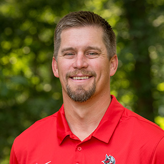 CJ Siewert, Director of Sports Information with short hair and a beard wearing a red collared shirt stands outdoors with trees in the background.