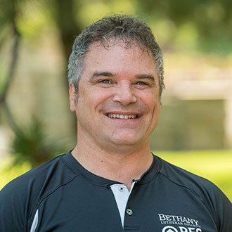 A man with short, curly gray hair is smiling outdoors with greenery in the background. He is wearing a dark Bethany Lutheran REC shirt.