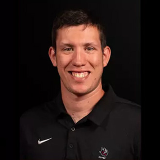 Dominic Santina, Head Coach – Cross Country/Track & Field wearing a black collared shirt with a Bethany Lutheran College logo smiles against a plain black background.