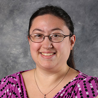 Gabrielle Emmons, Adjunct Faculty in the History Department and Online Instructor for the Academic Affairs Department, smiles in front of a gray mottled background.