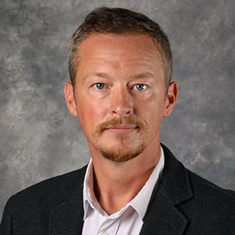 Will Frame, Adjunct Faculty and Lessons Instructor for the Music Department, stands against a gray background.