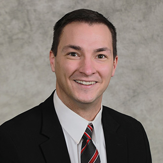 Pat Garvin, Head Men's Basketball Coach in a black suit, white shirt, and striped tie smiling in front of a gray textured background.