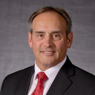 A middle-aged man in a dark suit, white shirt, and red patterned tie poses in front of a gray backdrop. If the context is Bethany Lutheran College and the file name indicates an official portrait or title, please provide it for a more specific caption.
