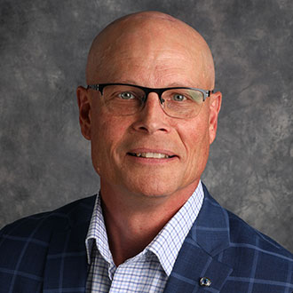 Bruce Gratz, Vice President of Institutional Advancement of the Advancement Department, poses in front of a gray background.