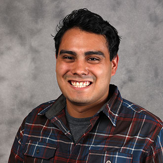 Bethany Lutheran College student with dark hair wearing a plaid shirt smiles at the camera in front of a gray textured background.