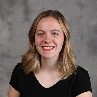 A Bethany Lutheran College student with shoulder-length blonde hair smiles in front of a gray studio backdrop, wearing a black shirt.
