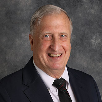 Robert Hanna, Professor for the English Department, smiles while posing in front of a gray textured background.