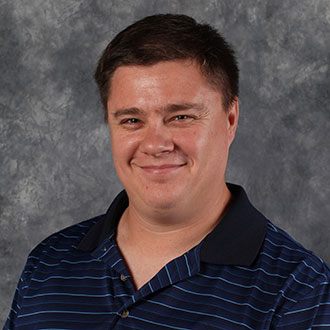 Benji Inniger, Adjunct Faculty for the Theatre Department, standing in front of a gray mottled background.